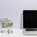 macbook pro on white table beside a miniature shopping cart with money