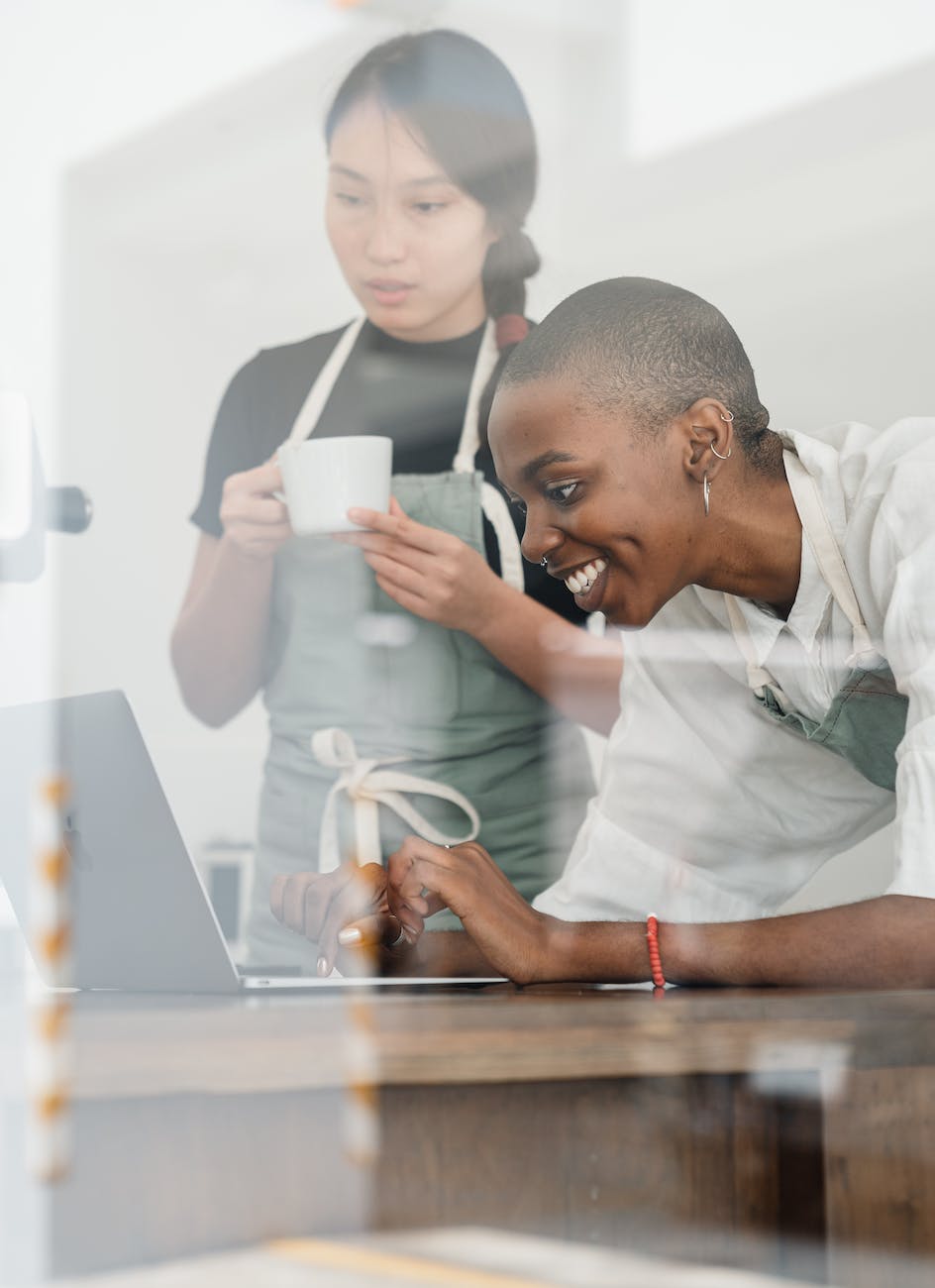 multiethnic female baristas in aprons using laptop during coffee break